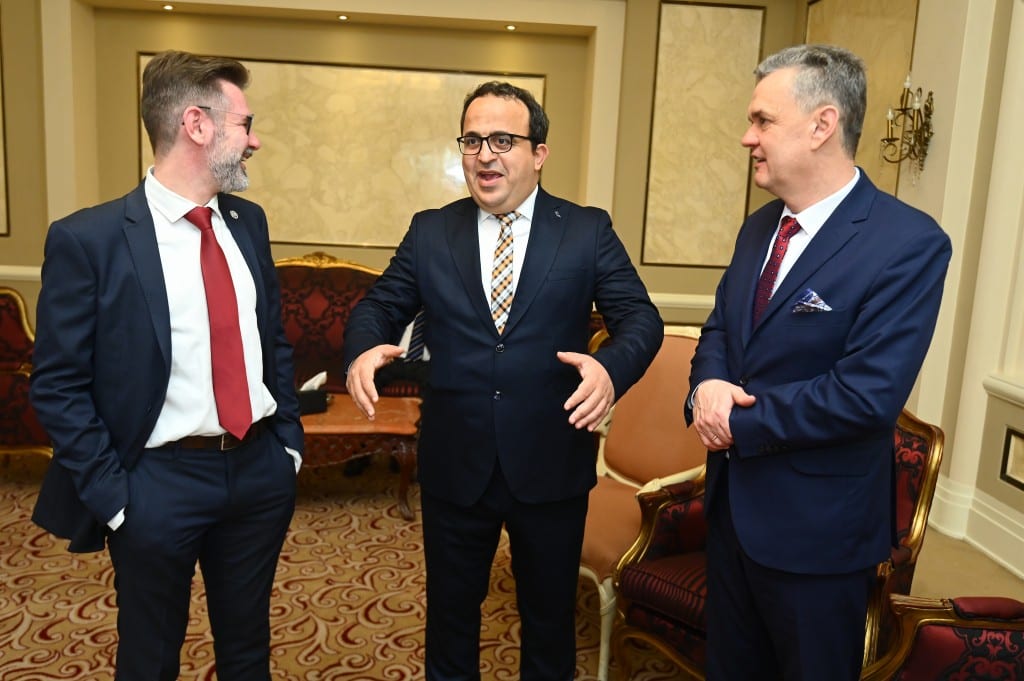 Three delegates in dark suits talk and laugh together in a hotel lounge with gilded armchairs.