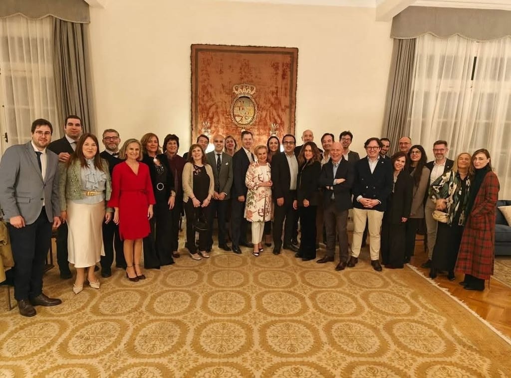 Delegates in formal dress gather for a group photograph in a reception room, beneath a large tapestry bearing a coat of arms.
