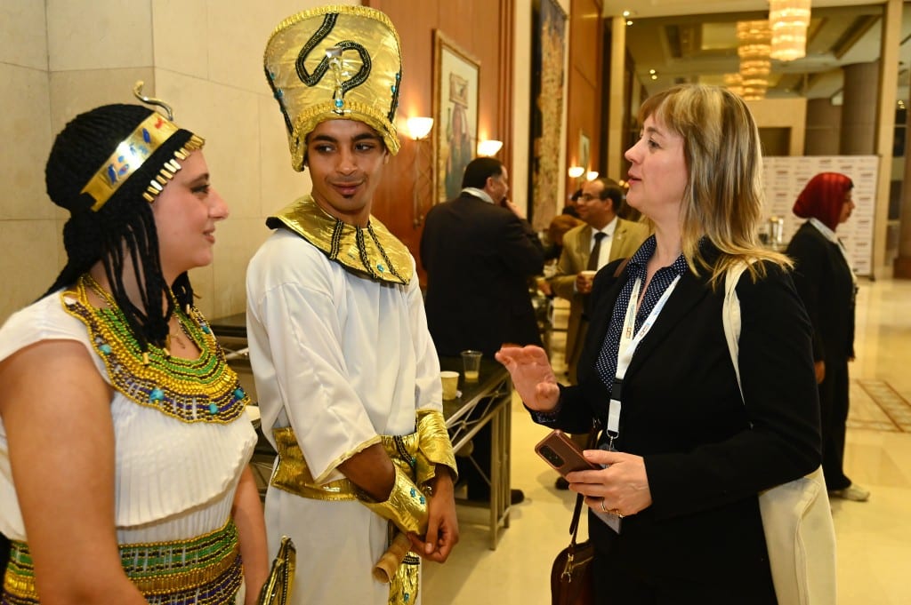 A host dressed as a pharaoh and another dressed as Cleopatra talk with a smiling delegate in a hotel lobby.